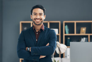 A person with their face obscured, standing confidently in an office setting, wearing a dark blazer and a watch.